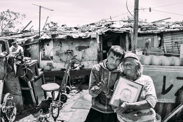 ‘Old’ Selva stands with her grandson and a picture of her own grandmother in front of her home (Patrick O'Hare, 2015)